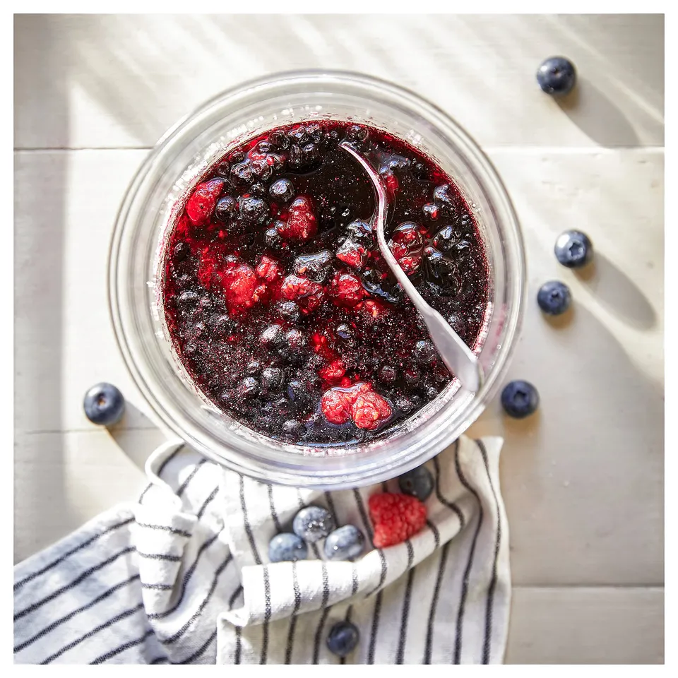Glass jar with mixed berry jam, spoon, scattered berries on cloth. Fresh, homemade look.