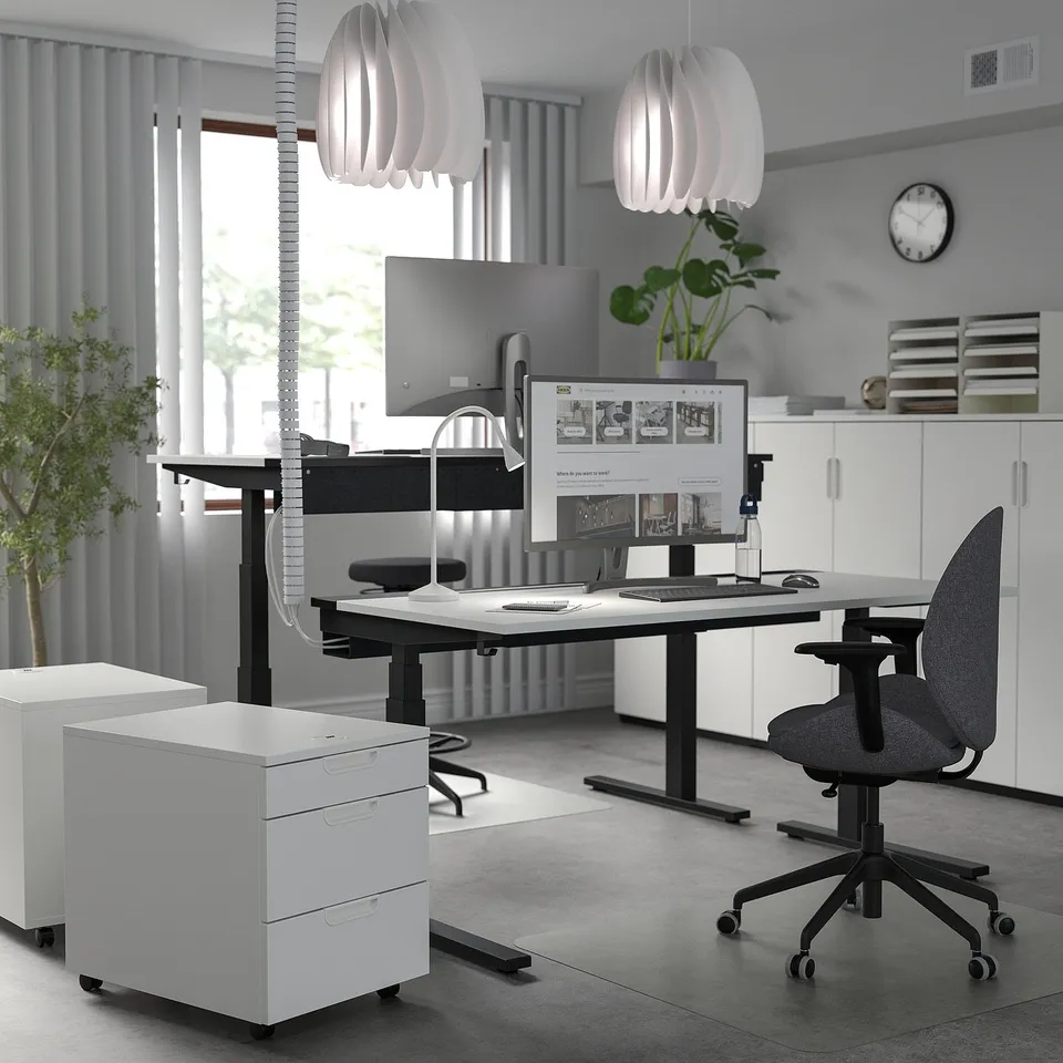 Modern minimalist office: MITTZON white sit-stand desk, sleek design, large screen, grey chair, white cabinets, plants, pendant lights.