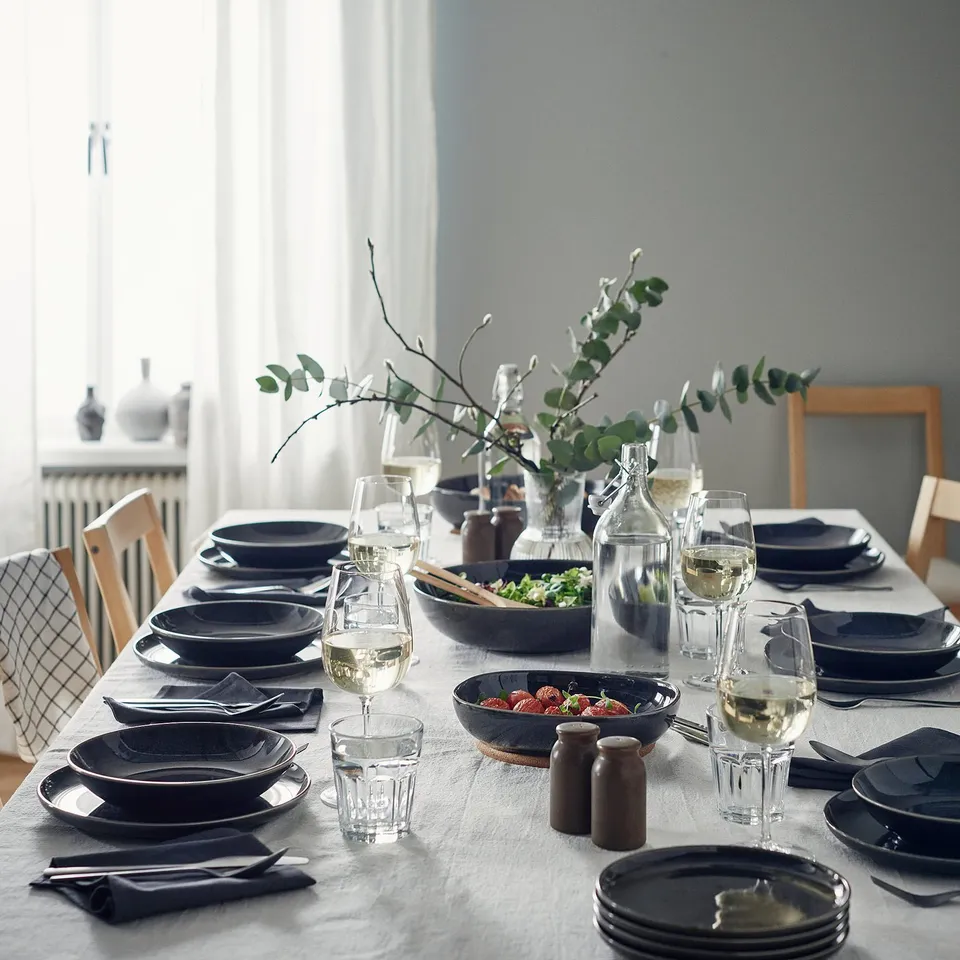 Table set with dark plates, glasses, cutlery, strawberries, salad, carafe, salt & pepper, flowers. White cloth, natural light.