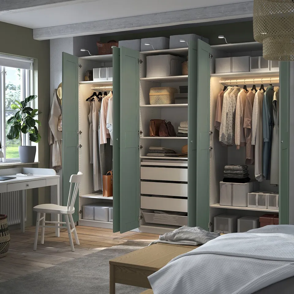 Bedroom with open green PAX wardrobe showing clothes, drawers, and storage boxes. White chair by window with plant. Bed partially visible.