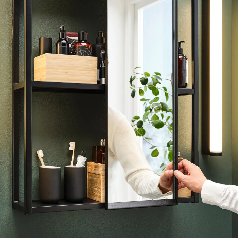 Person adjusts wall-mounted mirror, revealing shelving with toiletries and plant, against dark green backdrop in minimalist shelter.