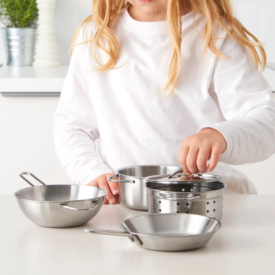 A girl in a white shirt plays with stainless steel toy cookware, holding a pots lid.