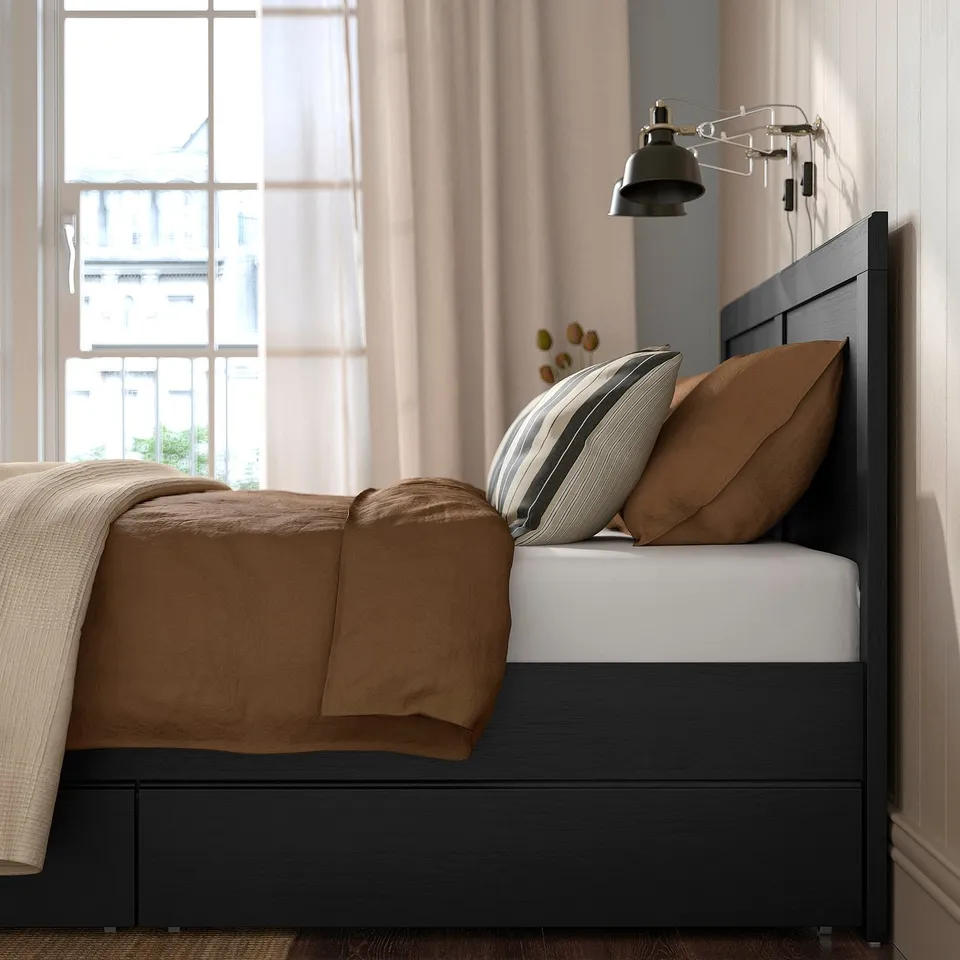 Bedroom scene with FÅGELFJÄLLET bed. Brown blankets, striped pillows. Drawers beneath bed, wall lamp above.