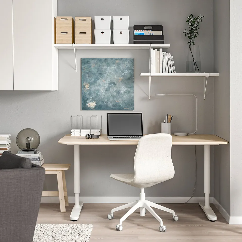Home office setup with white BERGSHULT desk, laptop, chair, shelves holding boxes, books, and a blue wall art.