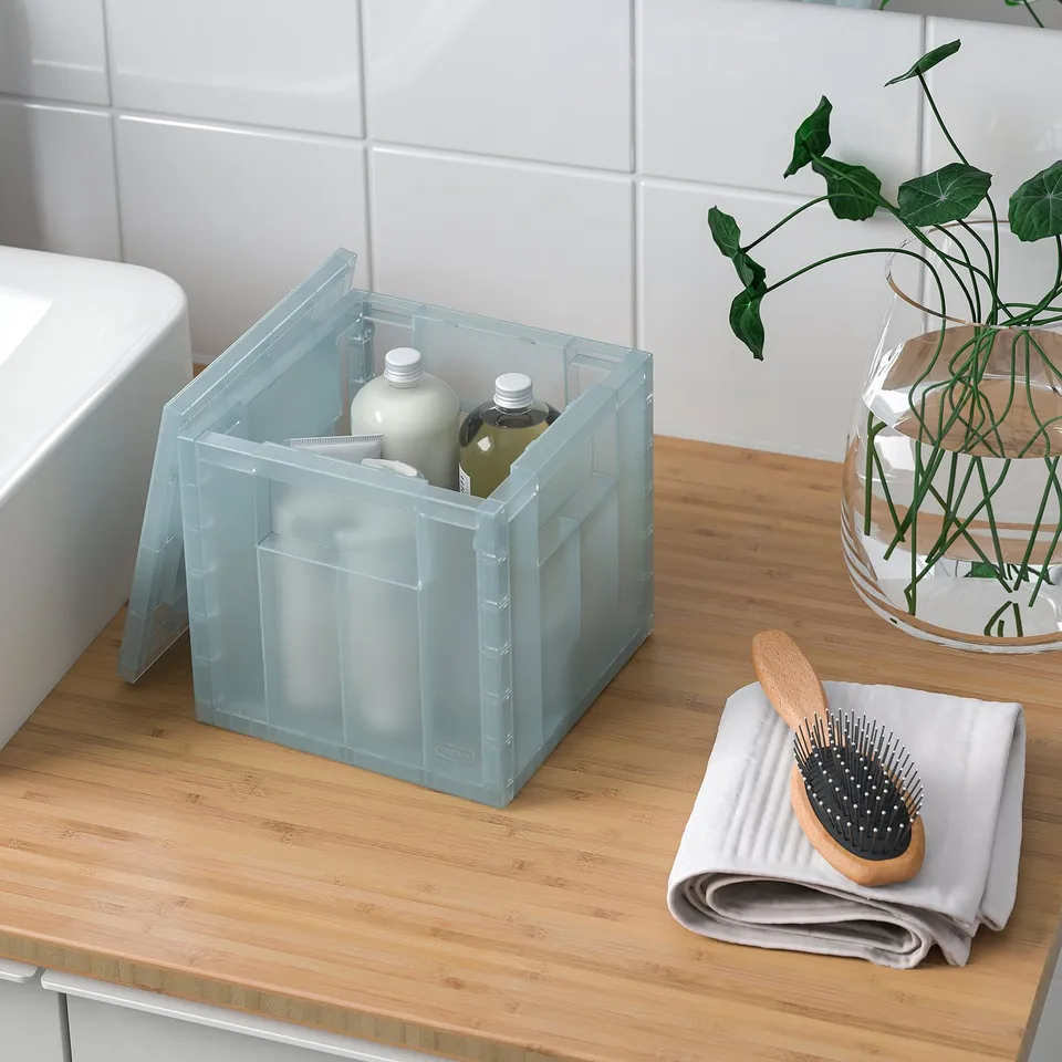 Clear plastic storage box with lid on a bathroom counter, holding bottles. Beside it, a folded towel and brush.