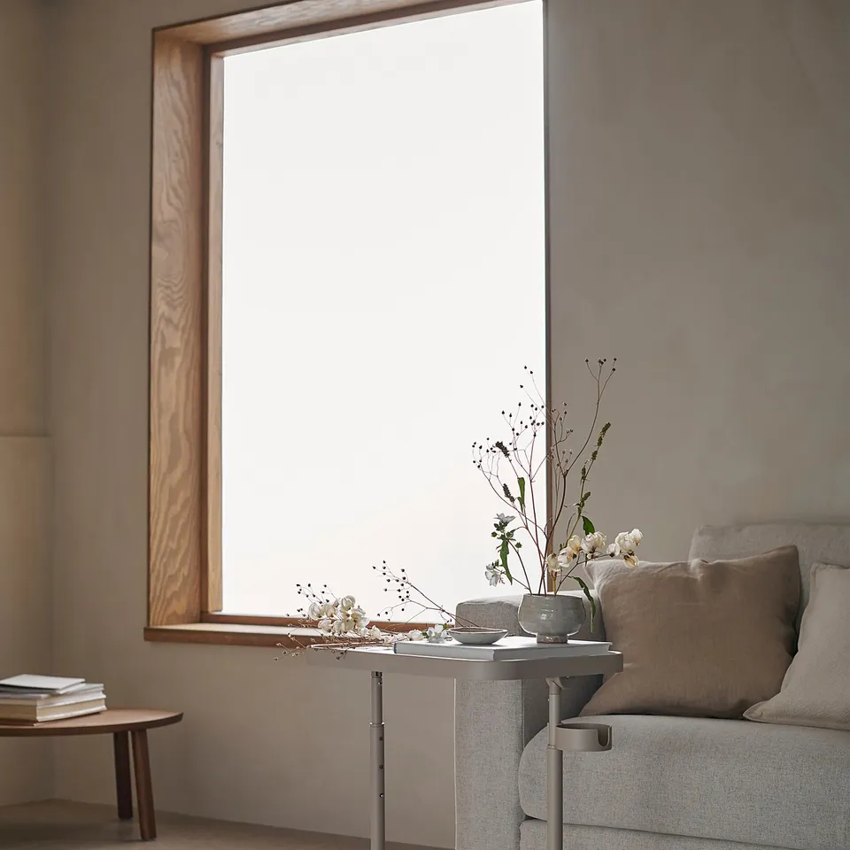 Modern living room with grey BJÖRKÅSEN laptop stand, cushioned chair, vase with flowers on a small table, and a large window.