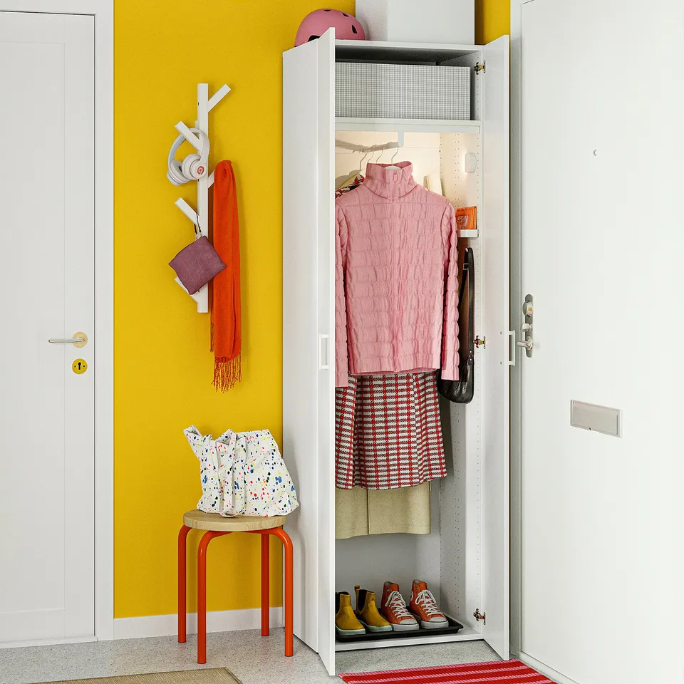 A small white closet with an open door revealing clothes and shoes organised inside, with colourful accessories hanging on the wall beside it.
