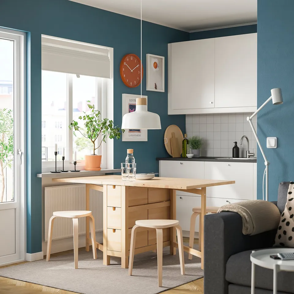 Modern kitchen with light wood table & stools, white cabinets, grey walls & large window.