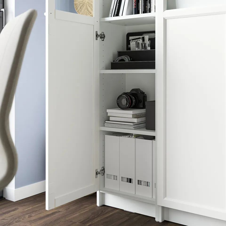 A white BILLY bookcase with glass panel; adjustable shelves hold books and a camera, while a cabinet below conceals clutter.