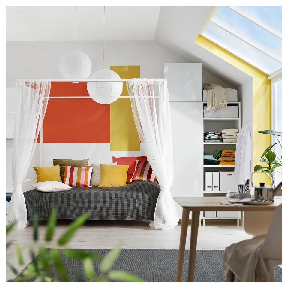 Modern bedroom with VITARNA bed, colourful cushions, white curtains, yellow accent wall, shelves with clothes & books, wooden chair.