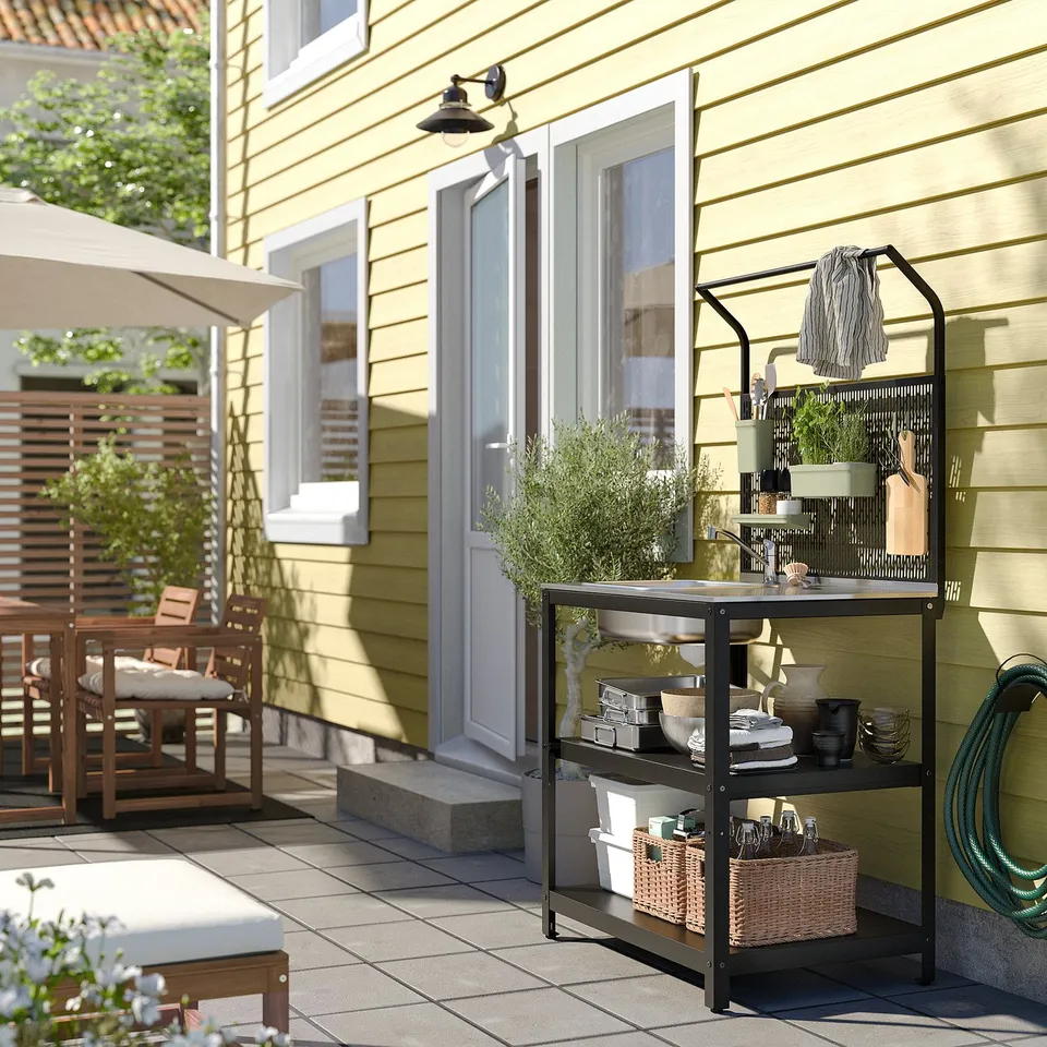 Modern outdoor kitchen setup: black metal sink stand with stainless steel basin, wooden dining area, yellow house.