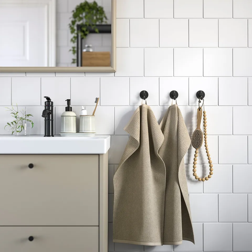 Modern bathroom with white tiles, beige cabinet, GULLDYNA towel, recycle cotton, black hooks, ceramic containers, and small plant.