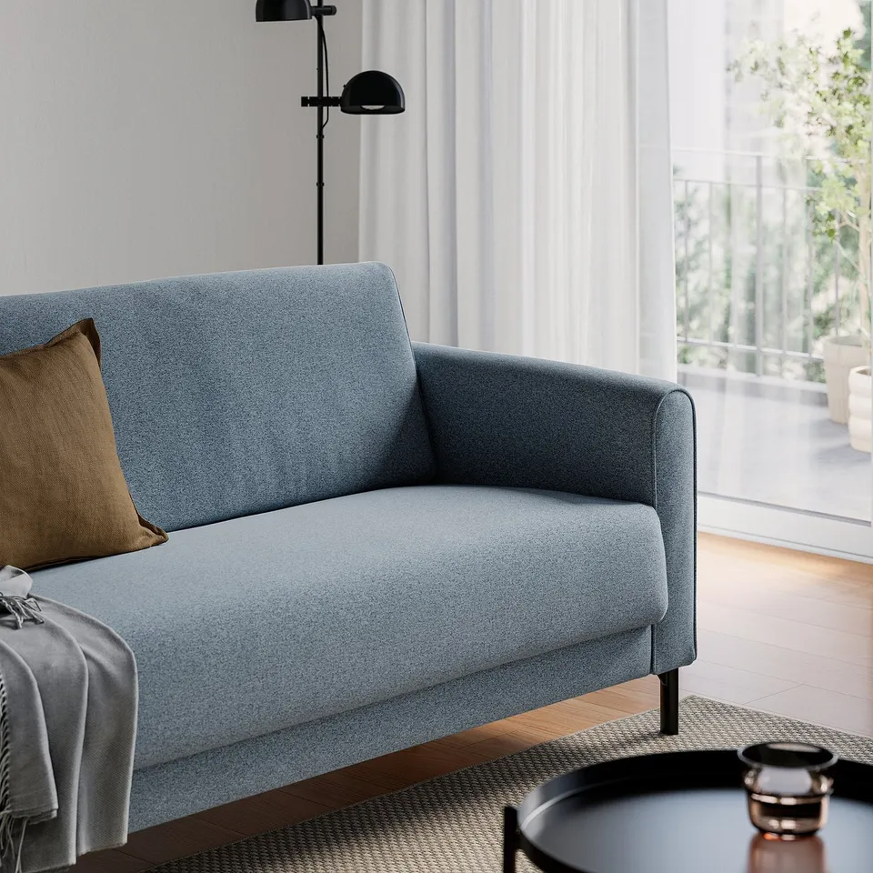 A cosy living room with a blue sofa, featuring a pillow, throw, and lamp, with large windows allowing natural light.