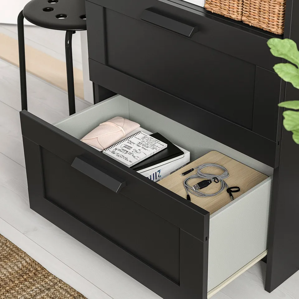 Modern desk with adjustable shelves, open drawer showing books, cables, and folded cloth, black and light wood finish.