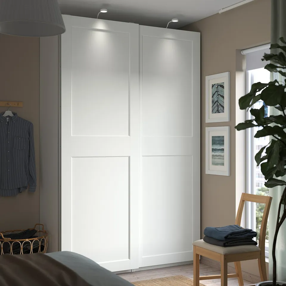 Modern bedroom with PAX wardrobe, sliding doors, and lighting. Clothes on wall rack, wooden chair, large plant by window. Nature prints on wall.