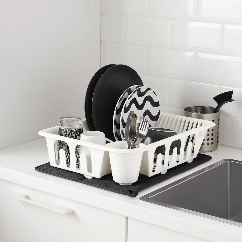 White sink area with items: a dish drainer holding plates, cups, and utensils, a drying mat underneath, and a brush nearby.