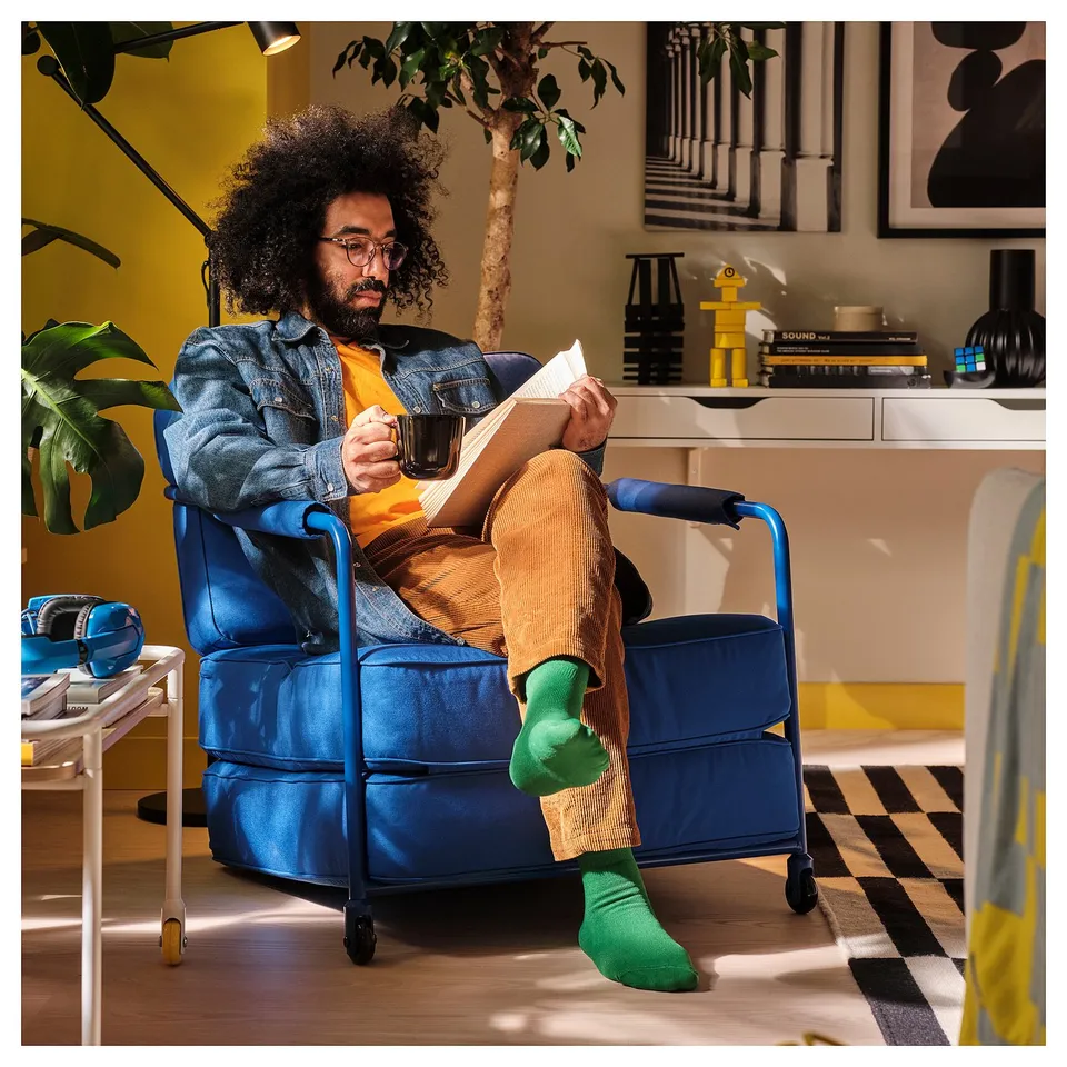 Person in blue BRÄNNBOLL chair, reading, holding mug. Chair has a folded seat cushion, castors for easy movement.