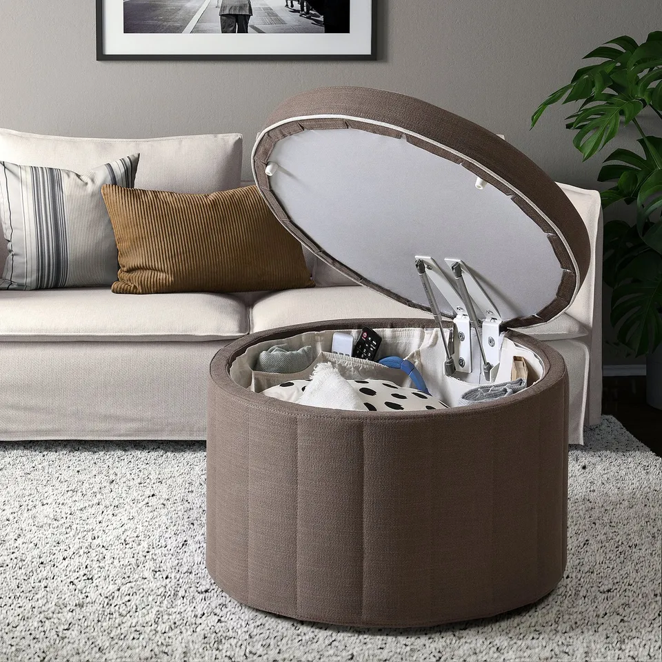 Lid-opened storage ottoman with inner pockets, revealing remote controls and blankets, beside a beige sofa on a white rug.