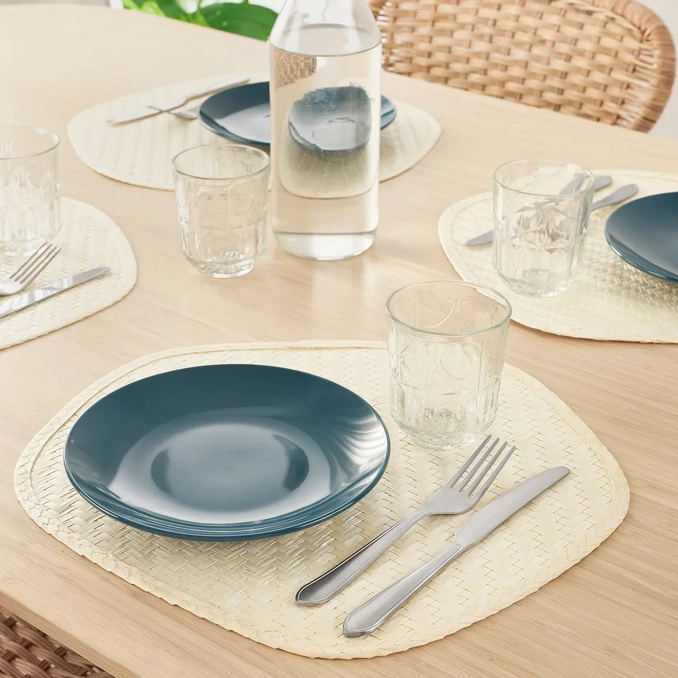 Table set with teal plates, clear glasses, silverware, on woven placemats. A bottle of liquid is in the centre.