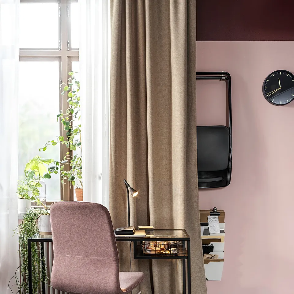 Cosy home office setup with pink ANNAKAJSA curtains, desk, chair, lamp, wall-mounted tv, clock, and plants. Dark floor, light walls.