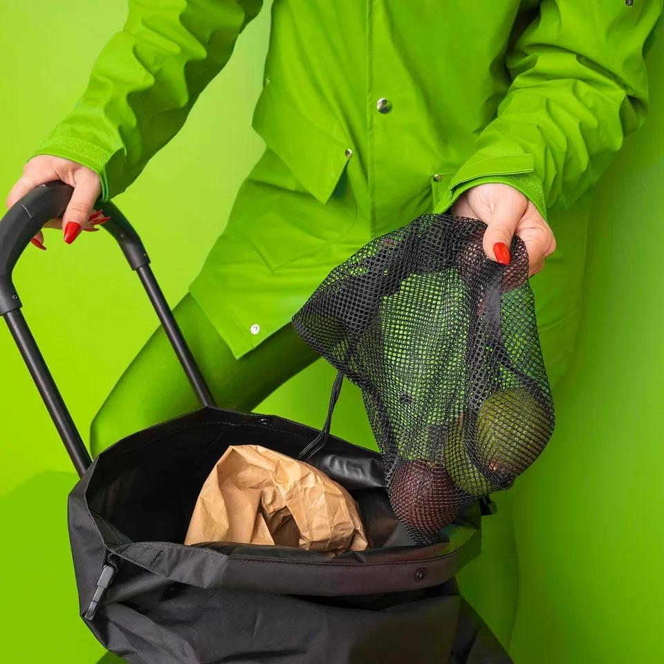 Person in neon green jacket holds mesh bag with groceries, pushing a black shopping cart against a green background.