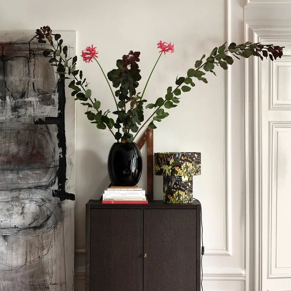 Dark wooden cabinet holds black vase with pink flowers, surrounded by art books and decorative box near a white door.
