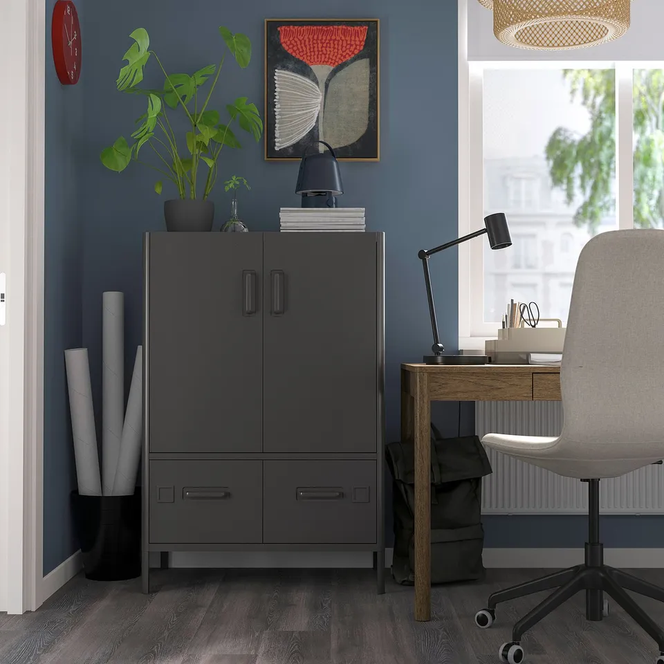 Modern home office with grey IDÅSEN cabinet, wooden desk, grey chair, blue wall, red clock, painting, black lamp, plant.