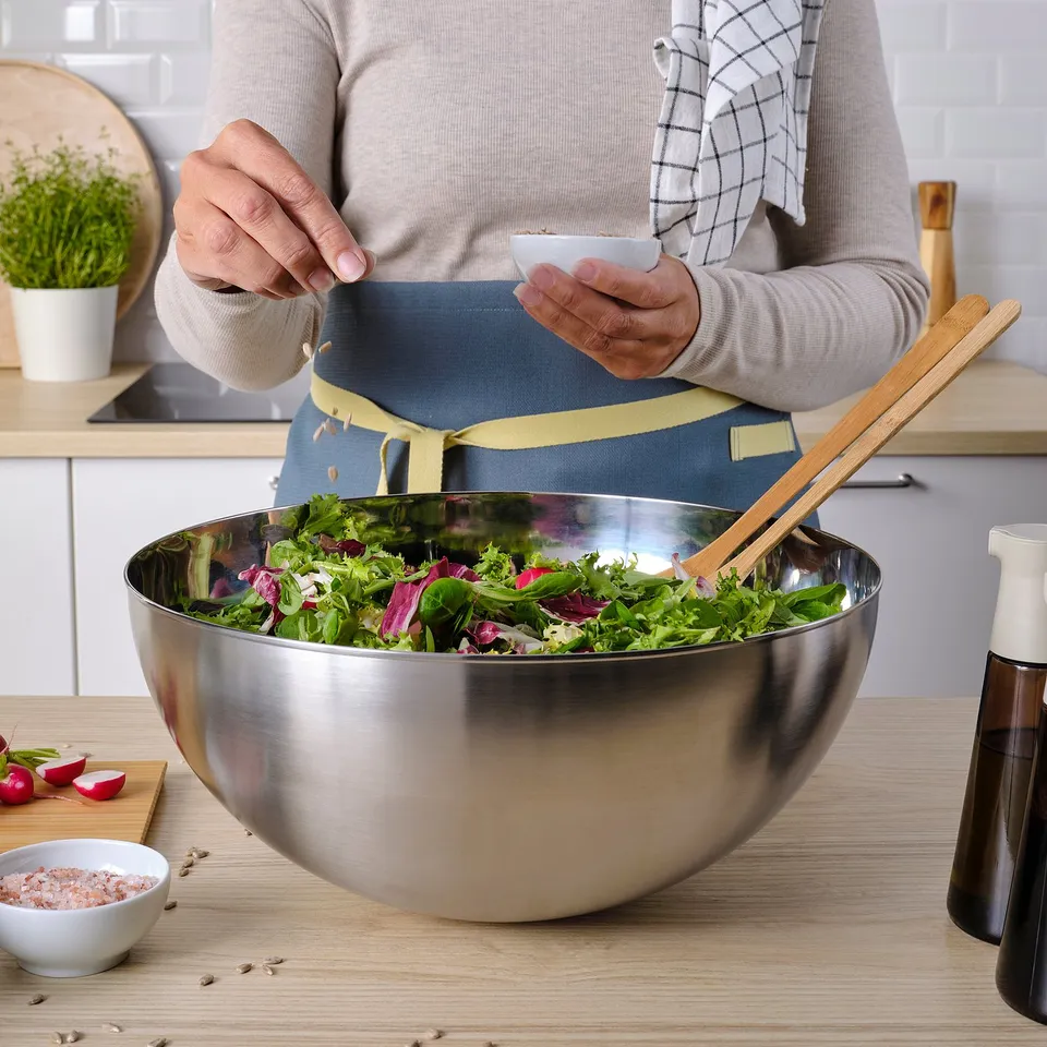 A person in an apron adds seeds to a large steel mixing bowl of salad vegetables on a kitchen countertop.