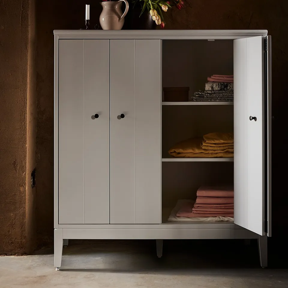 Modern light grey cabinet with open doors revealing folded textiles, featuring minimalist design with black handles, and a flower vase atop.