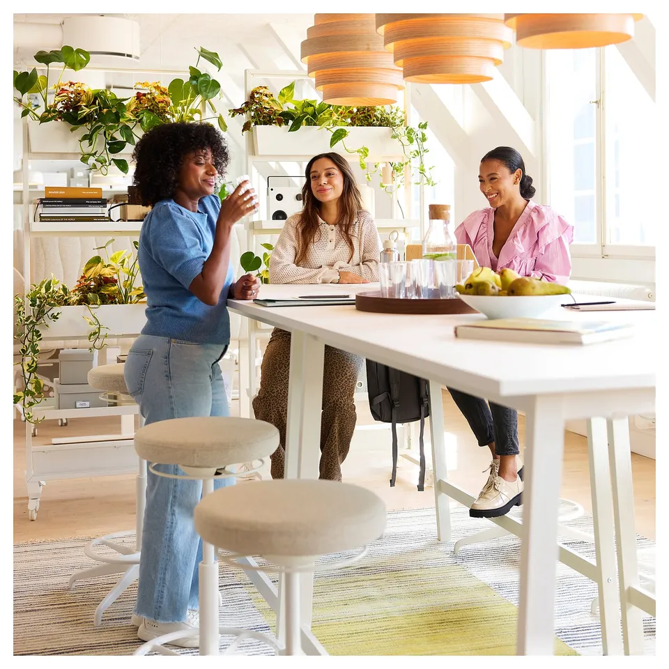 Three people collaborate at a modern MITTZON table with cable management, surrounded by soft stools, colourful rugs, and hanging plants.