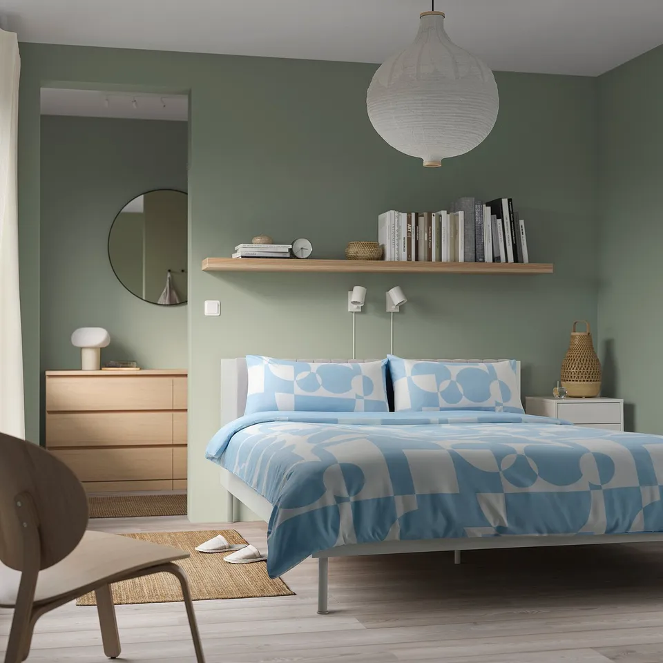 Modern bedroom with sage walls, white BRUKSVARA bed frame, blue circular bedding, wooden dresser, hanging light, bookshelf, and chair in corner.