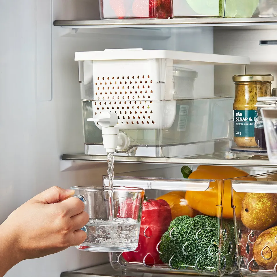 A hand fills a clear cup with water from a tap on a white fridge container, surrounded by fruits and veggies.