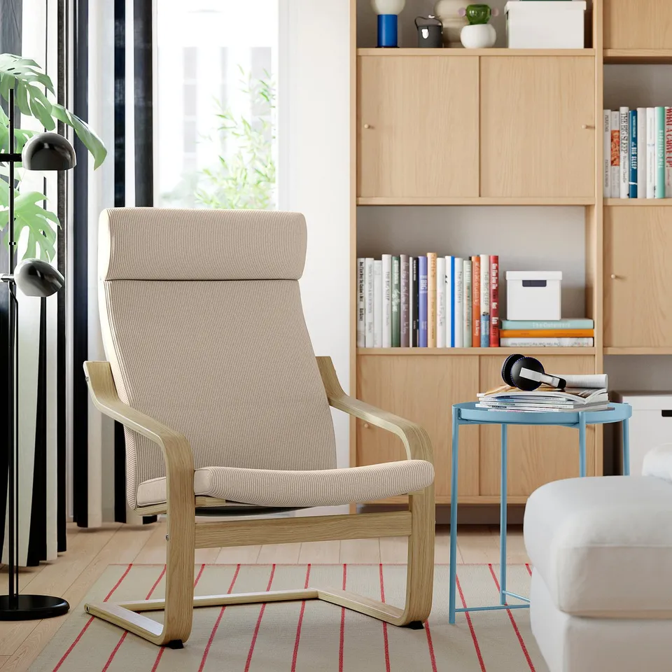 Beige IKEA POÄNG armchair on red rug, beside blue side table with books & headphones, set against a bookshelf in a cosy modern living room.