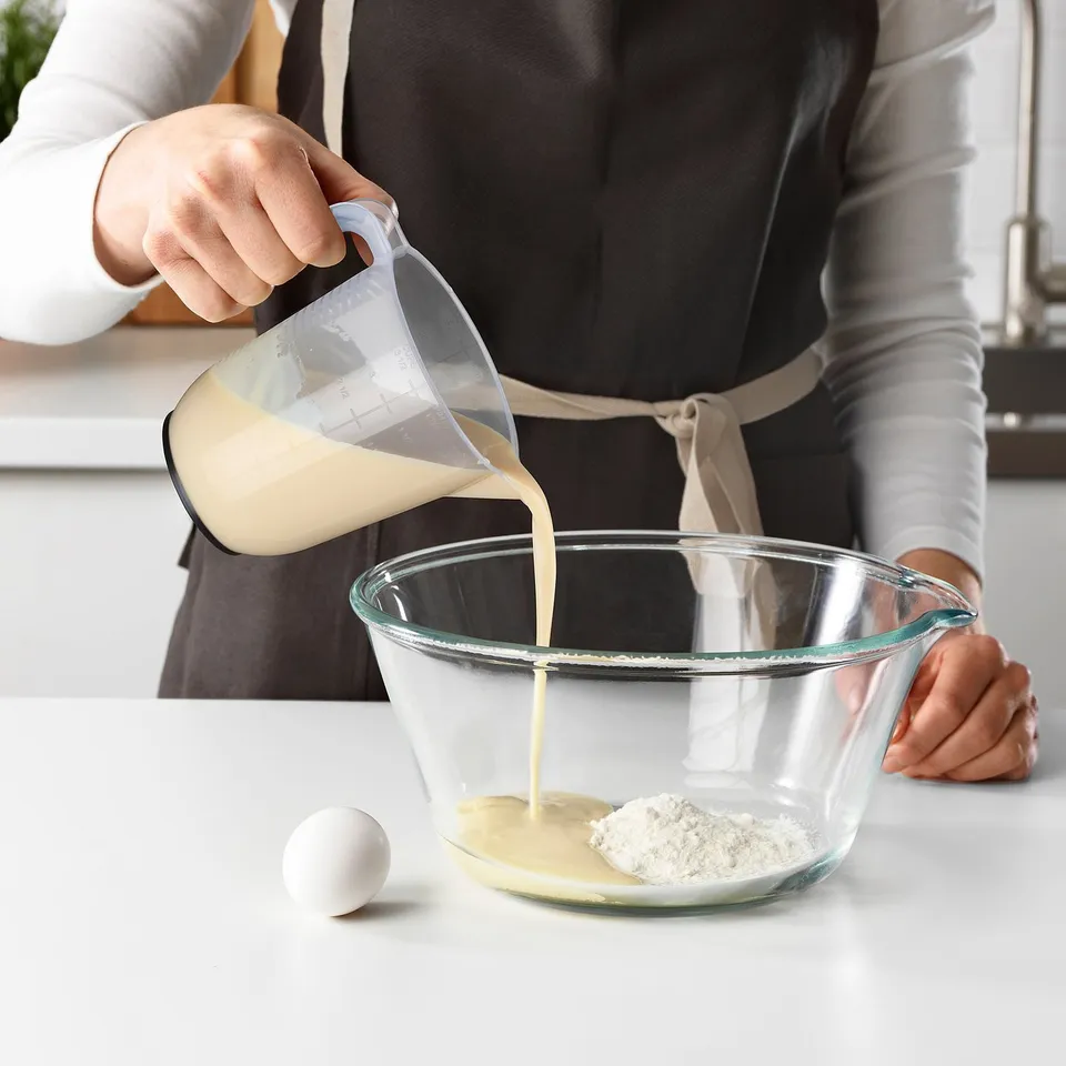 Person pouring liquid from a transparent measuring jug into a glass bowl on a kitchen counter. The jug has multiple measurement scales.