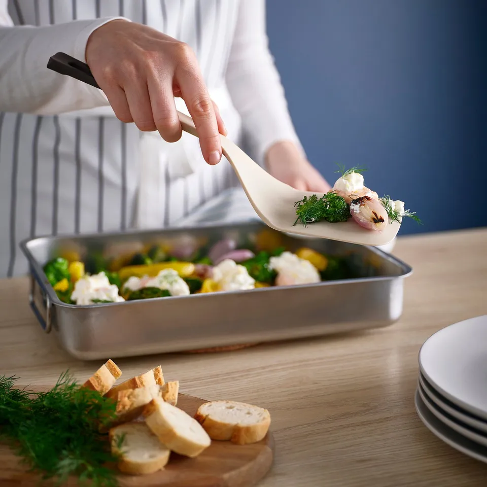 A person uses a colourful spatula to serve food from a metal dish. The spatulas wide blade and sloped handle help easily flip and portion dishes.