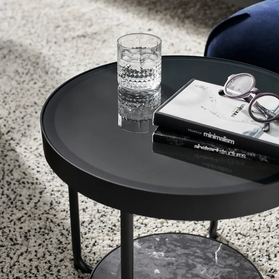 Black round side table with glass top, holding a glass and books, with sleek metal legs.