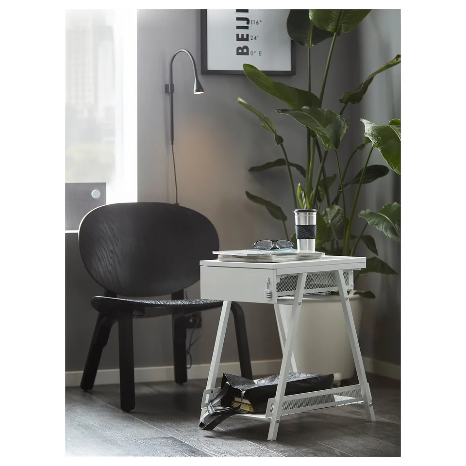 Modern black desk chair and white metal side table with drawer and lower shelf. Large green plant and wall-mounted lamp in background.