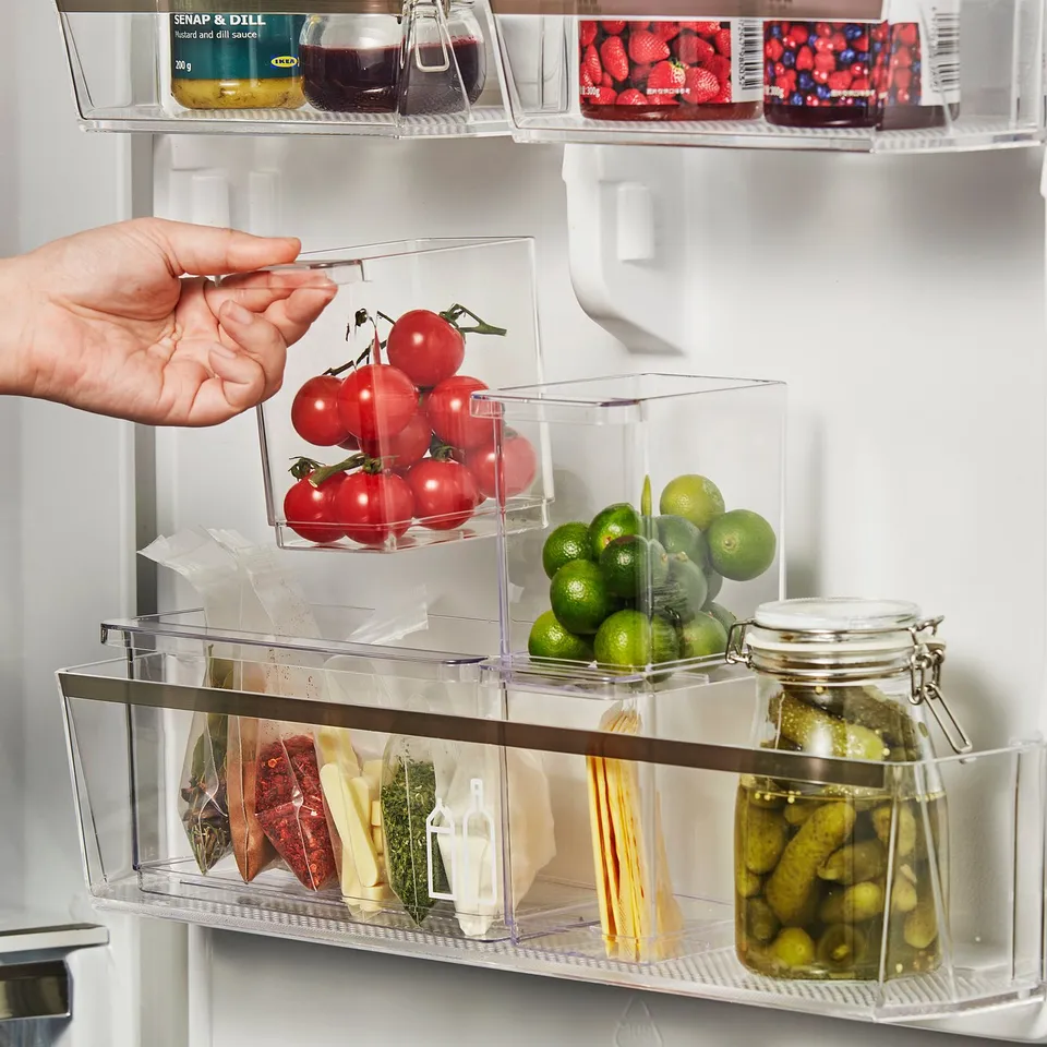 Hand reaching into fridge with clear plastic storage boxes containing tomatoes, limes, cheese, and pickles. The boxes have handles for easy lifting.