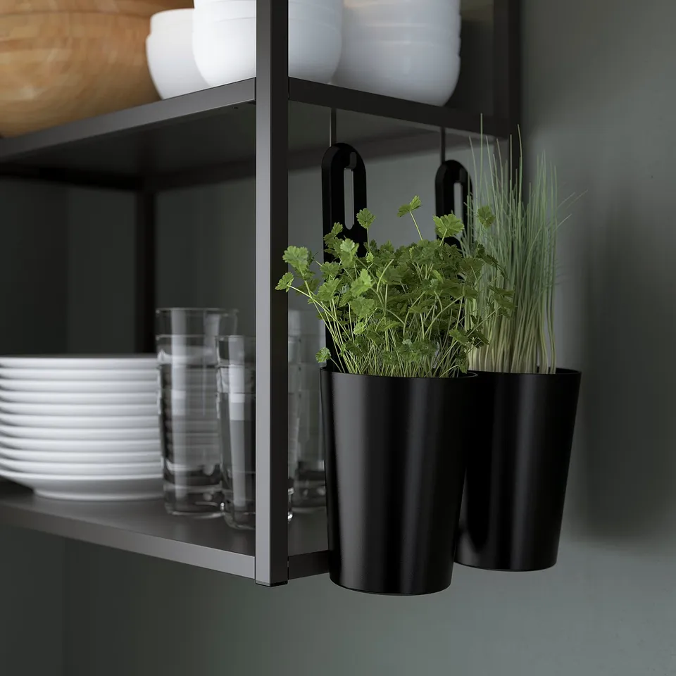 Black kitchen rail with hooks holding pots, one with fresh herbs, under a shelf with glasses and bowls.