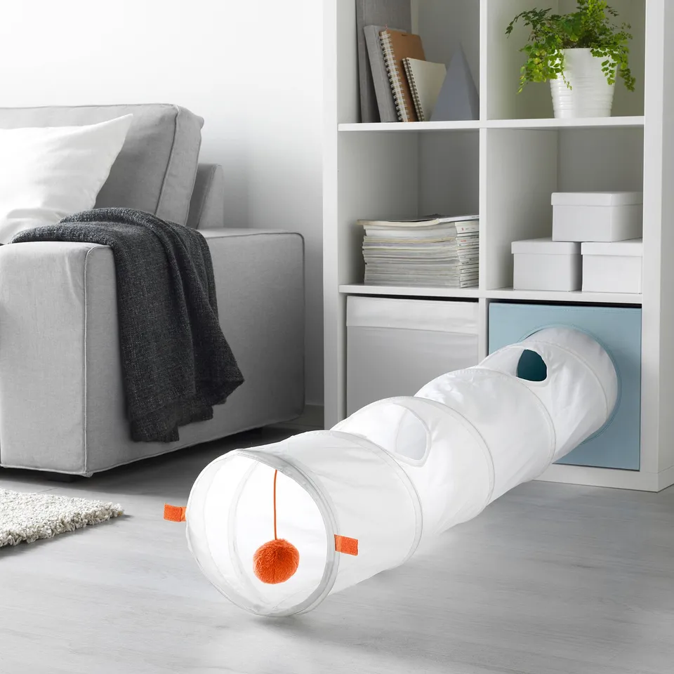 A cat play tunnel with a dangling orange pom-pom toy, set near a white couch and shelves.