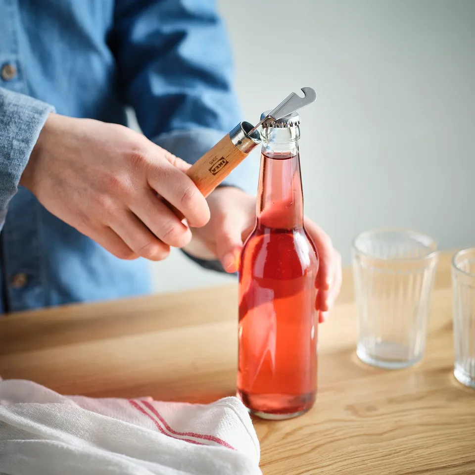 Person opens a red drink bottle using a dual bottle/can opener with a wooden handle on a wooden table.