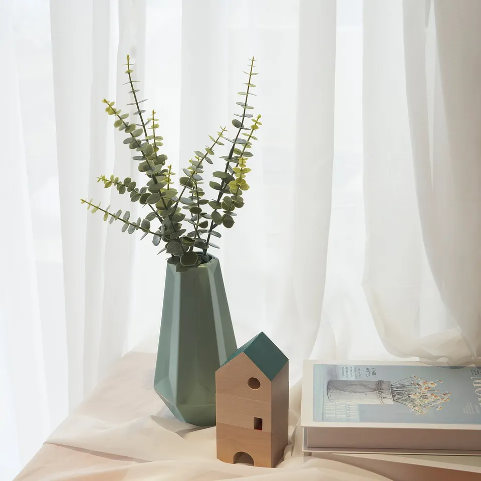 A teal vase with eucalyptus branches sits on a white shelf next to a wooden house-shaped planter and a book.
