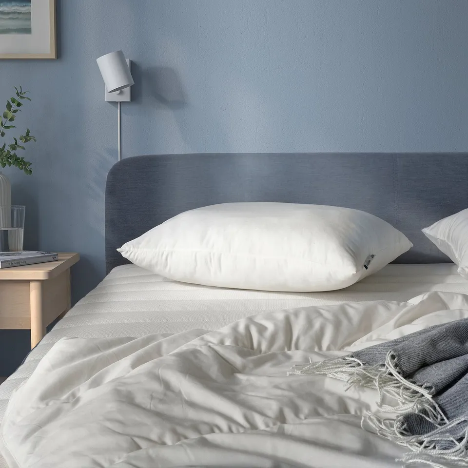 Cosy bedroom with LAPPTÅTEL pillow on bed, grey headboard. White linens, bedside table with lamp, plant, books, and vases.