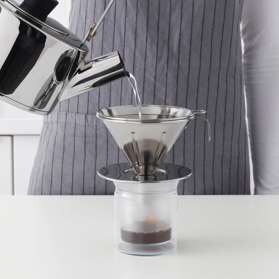 A person pours water from a kettle into a metal coffee philtre placed on a glass jar. The philtre allows oils, enhancing coffee flavour.