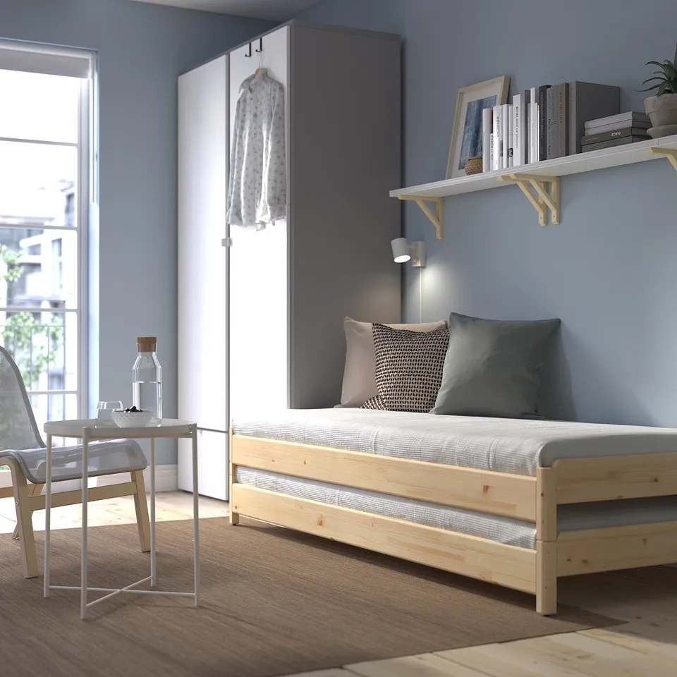 Minimalist bedroom with UTÅKER light pine stackable bed, grey pillows, bleached oak floor, and large window; shelf with books and cloth hanger beside.