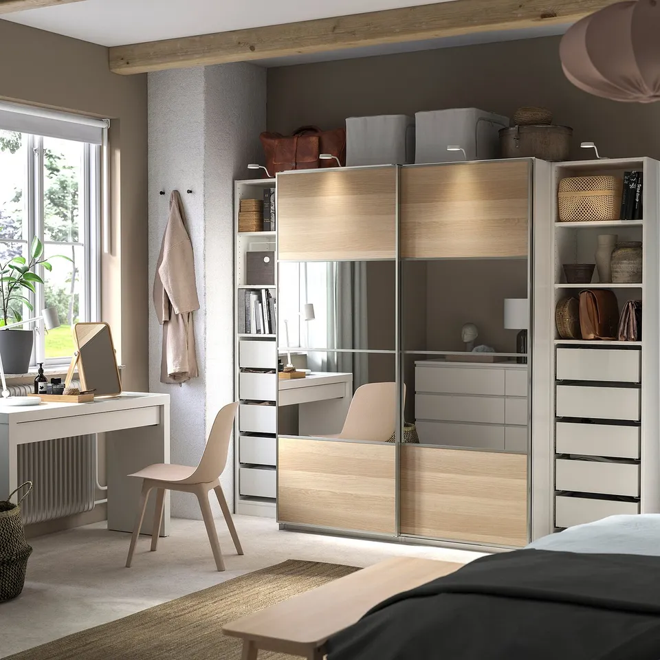 Modern bedroom with a beige PAX wardrobe, featuring mirrored sliding doors, various shelves, and drawers. It includes a desk, chair, and accessories.