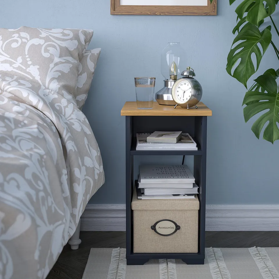 Bedside table SKRUVBY next to a bed. Silver clock, books, and plant on top. Blue wall and striped rug below.