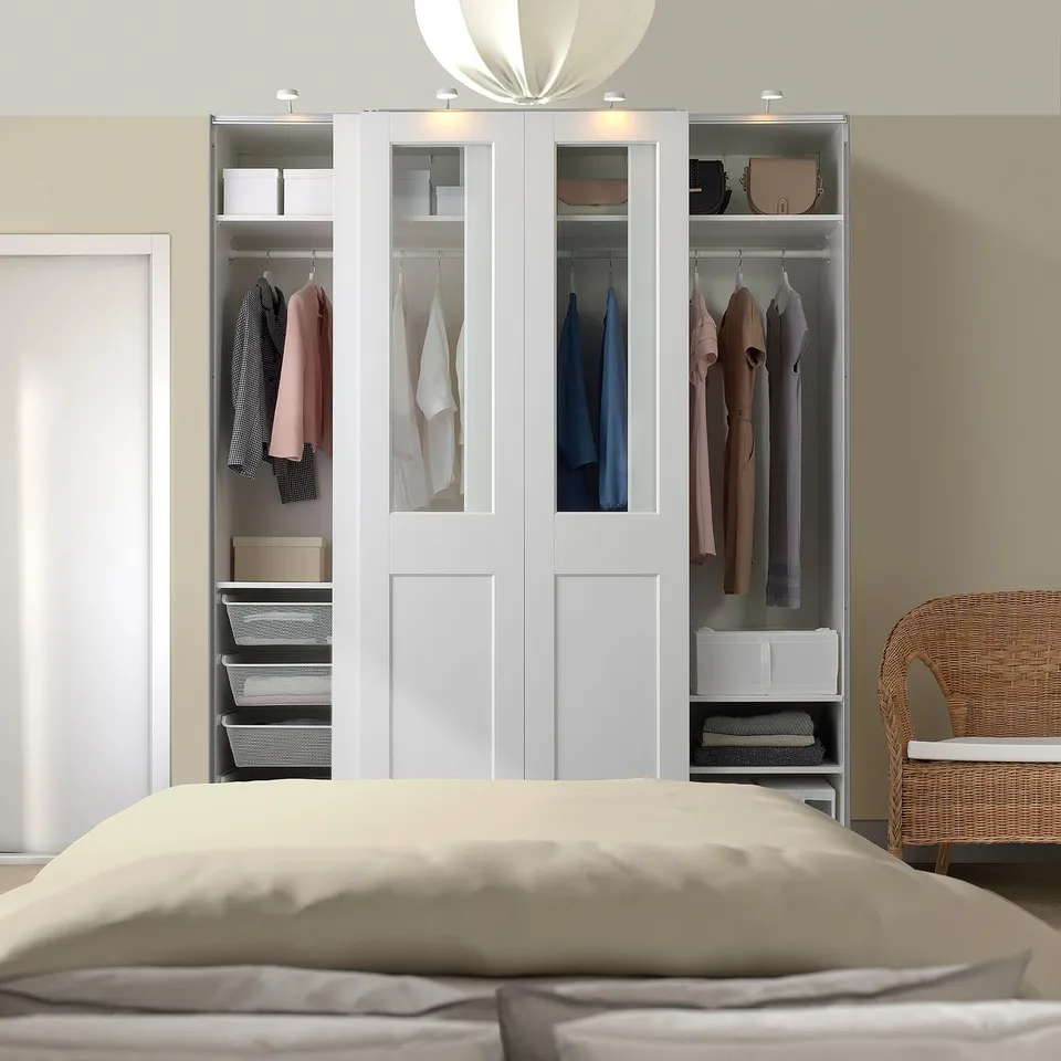 A large white PAX wardrobe with glass, displaying clothes and organised items. It stands tall beside a wicker chair and bed with beige bedding.