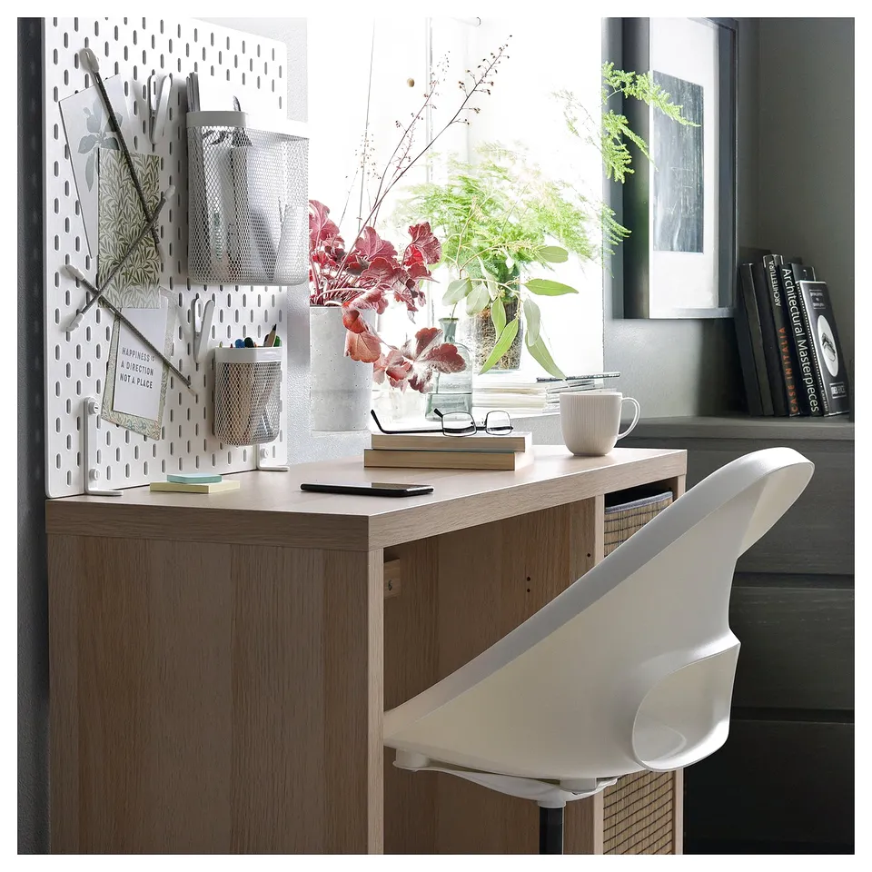 Modern home office setup with wooden desk, white chair, and wall organiser. Decorated with flowers and books.