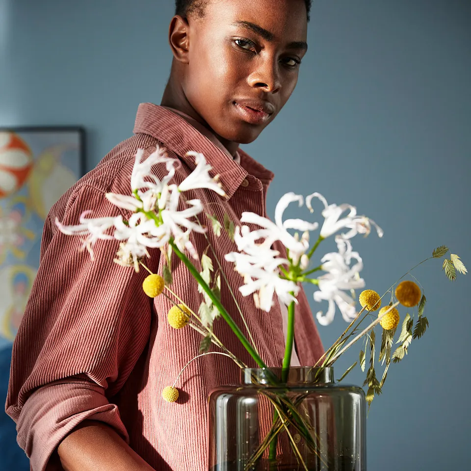 A person wearing a red shirt holds a transparent brown glass vase with white flowers.